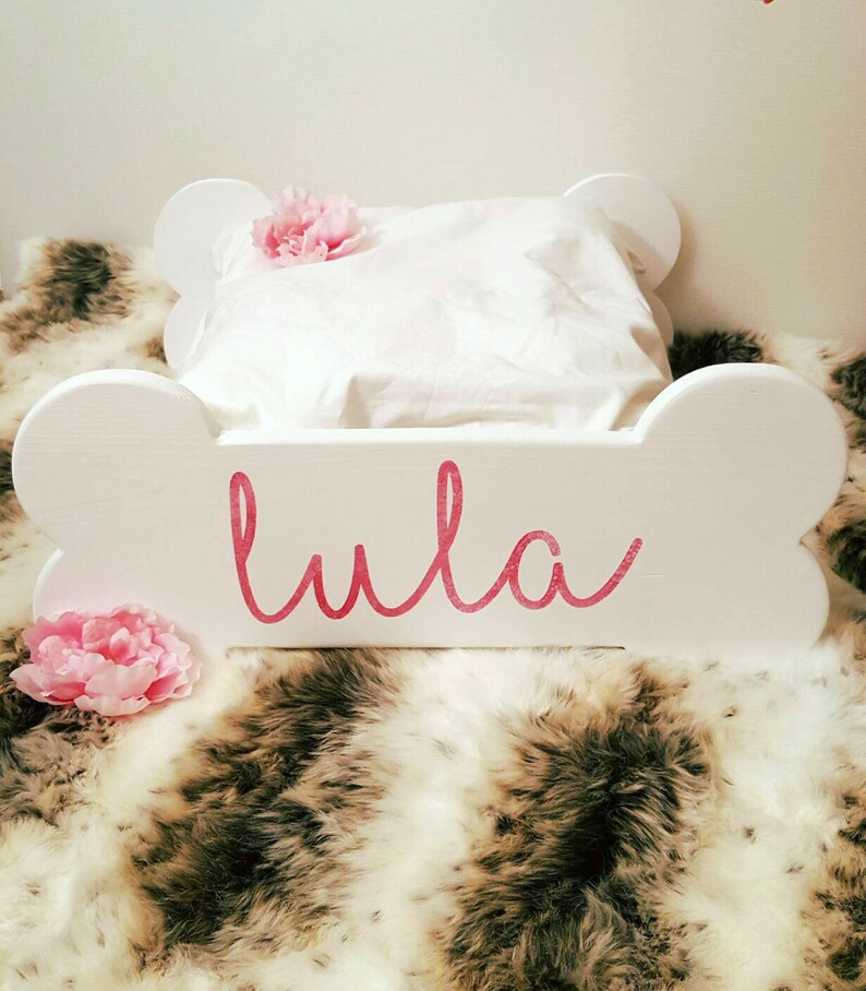 May include: A white, bone-shaped dog bed with the name "Lula" in pink script. It features a white pillow and is adorned with pink flowers. The bed rests on a faux fur blanket.