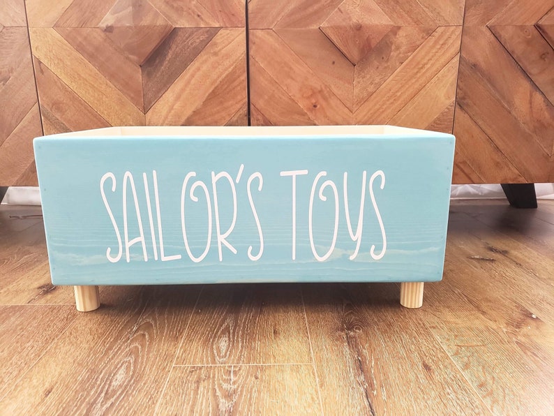 May include: A light blue wooden toy box with the words "SAILOR'S TOYS" written in white on the front. The box has four wooden legs.