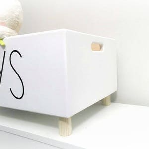 May include: White wooden toy storage box with the word "TOYS" printed in black on the front. The box has a wooden handle and sits on four wooden legs.