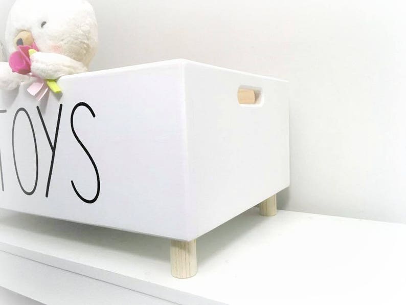May include: White wooden toy storage box with the word "TOYS" printed in black on the front. The box has a wooden handle and sits on four wooden legs.