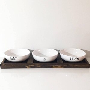 May include: Three white ceramic pet bowls with the names "Max", "Ace", and "Luna" printed on them, sitting on a dark brown wooden tray.