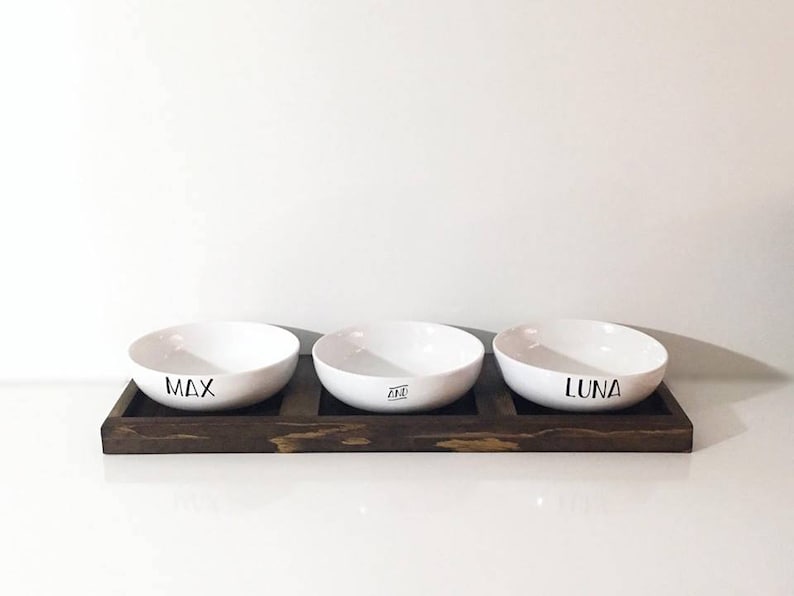 May include: Three white ceramic pet bowls with the names "Max", "Ace", and "Luna" printed on them, sitting on a dark brown wooden tray.