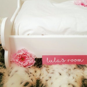 May include: A white bed frame with a white pillow and a pink flower. A pink sign that says "lula's room" is in front of the bed. The bed is on a faux fur rug.