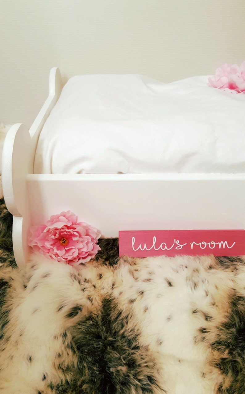 May include: A white bed frame with a white pillow and a pink flower. A pink sign that says "lula's room" is in front of the bed. The bed is on a faux fur rug.