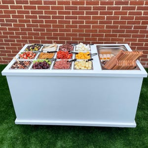 Puede incluir: Un carro de servicio blanco con m&uacute;ltiples recipientes de acero inoxidable llenos de varios alimentos, incluyendo fruta, queso, galletas y carne. El carro tiene una encimera blanca y una base blanca con ruedas. Hay tablas de cortar de madera en el fregadero en el lado derecho del carro.