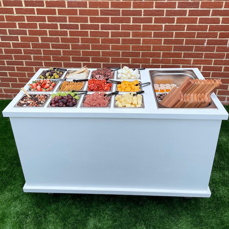 Puede incluir: Un carro de servicio blanco con m&uacute;ltiples recipientes de acero inoxidable llenos de varios alimentos, incluyendo fruta, queso, galletas y carne. El carro tiene una encimera blanca y una base blanca con ruedas. Hay tablas de cortar de madera en el fregadero en el lado derecho del carro.