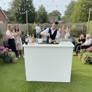 May include: A white bar in a garden setting, with a bartender preparing drinks. Various bottles and glasses are arranged on the bar. Guests are seen in the background, with decorative string lights and green grass.