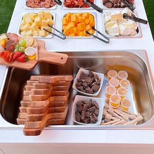 Puede incluir: Un carro de servicio blanco con una variedad de aperitivos y golosinas, que incluyen queso, galletas, fruta, chocolate y miel. El carro tiene un fregadero de acero inoxidable y una tabla de cortar de madera.