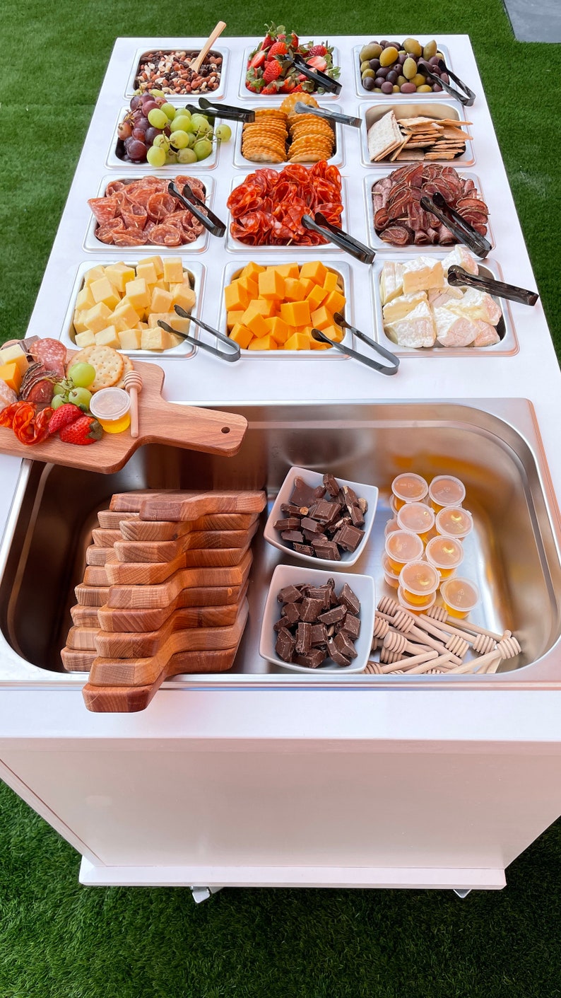 Puede incluir: Un carro de servicio blanco con una variedad de aperitivos y golosinas, que incluyen queso, galletas, fruta, chocolate y miel. El carro tiene un fregadero de acero inoxidable y una tabla de cortar de madera.