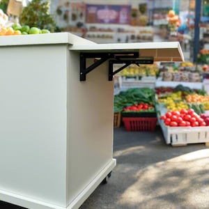 Puede incluir: Mesa plegable blanca con soportes de metal negro, sobre ruedas, en un mercado con productos frescos en el fondo.
