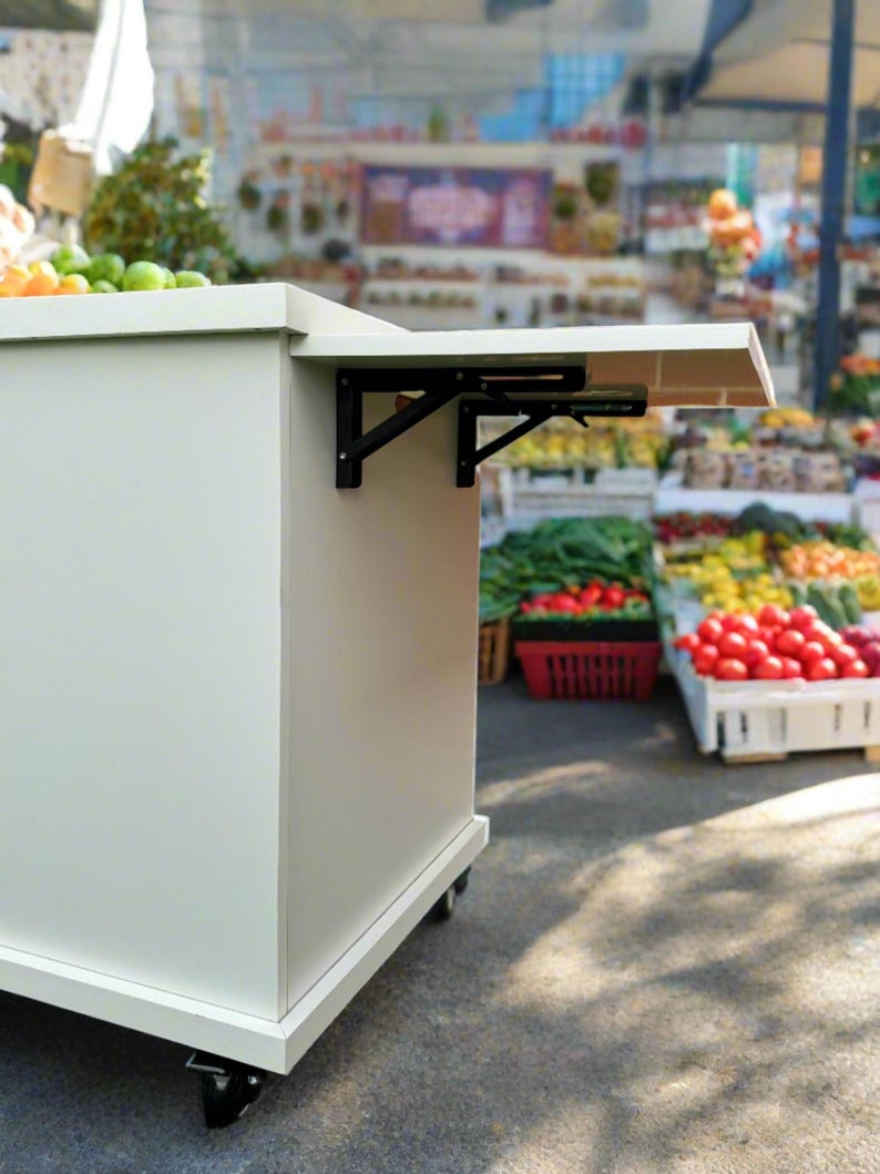 Puede incluir: Mesa plegable blanca con soportes de metal negro, sobre ruedas, en un mercado con productos frescos en el fondo.