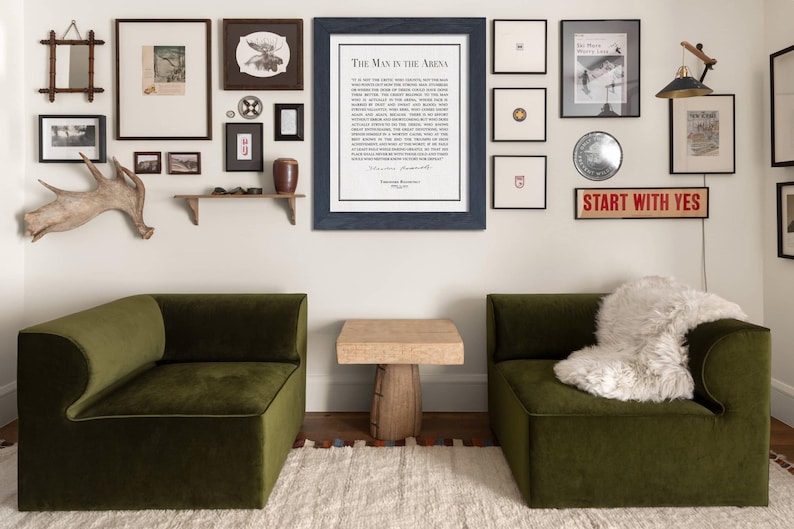 K&ouml;nnte beinhalten: Ein Innendesign-Szene mit einer Galeriewand mit gerahmter Kunst und einem Schild mit der Aufschrift "START WITH YES". Zwei olivgr&uuml;ne Samtsessel stehen sich gegen&uuml;ber, mit einem kleinen Holztisch dazwischen. Ein wei&szlig;er Teppich und ein dekoratives Geweih erg&auml;nzen die Einrichtung.