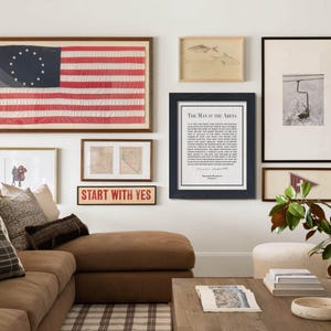 K&ouml;nnte beinhalten: Ein Wohnzimmer mit einem braunen Ecksofa, einem Holztisch und einer Bilderwand mit gerahmten Kunstwerken. Die Wand umfasst eine gerahmte amerikanische Flagge, Drucke und ein Schild mit der Aufschrift "START WITH YES".