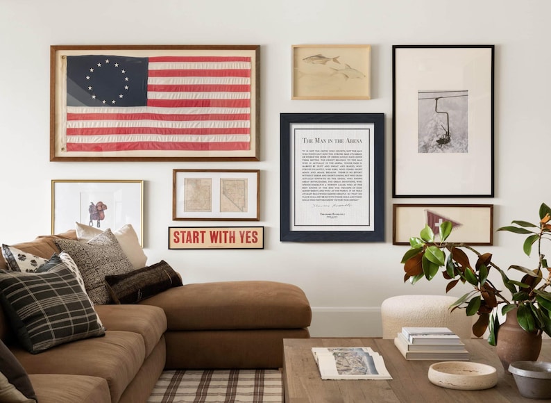K&ouml;nnte beinhalten: Ein Wohnzimmer mit einem braunen Ecksofa, einem Holztisch und einer Bilderwand mit gerahmten Kunstwerken. Die Wand umfasst eine gerahmte amerikanische Flagge, Drucke und ein Schild mit der Aufschrift "START WITH YES".