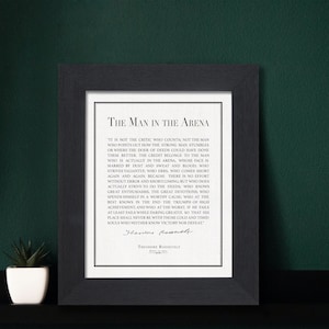 K&ouml;nnte beinhalten: Ein gerahmter Druck mit dem Text "The Man in the Arena" von Theodore Roosevelt. Der schwarze Rahmen umgibt einen wei&szlig;en Hintergrund mit dem Zitat in schwarzer Schrift. Links vom Rahmen steht eine kleine Topfpflanze.