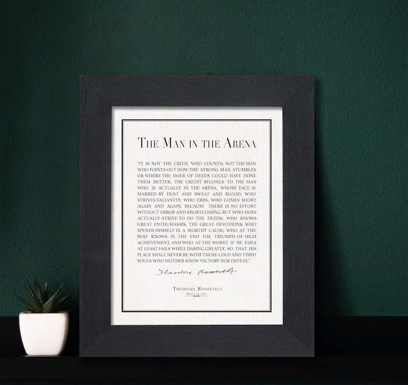K&ouml;nnte beinhalten: Ein gerahmter Druck mit dem Text "The Man in the Arena" von Theodore Roosevelt. Der schwarze Rahmen umgibt einen wei&szlig;en Hintergrund mit dem Zitat in schwarzer Schrift. Links vom Rahmen steht eine kleine Topfpflanze.