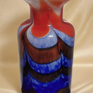 Op de afbeelding: Een decoratieve glazen vaas met een uitlopende bovenkant. De vaas heeft een wervelend patroon van rode, witte, blauwe en bruine kleuren. De vaas is cilindrisch met een brede basis en een smalle hals.