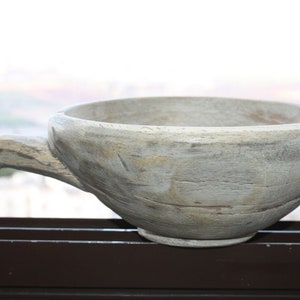 Op de afbeelding: Een rustieke houten kom met een handvat. De kom is lichtgrijs met zichtbare houtnerf en een afgeronde vorm. Het handvat steekt uit de zijkant, wat een uniek ontwerp creëert. Ideaal om te serveren of tentoon te stellen.