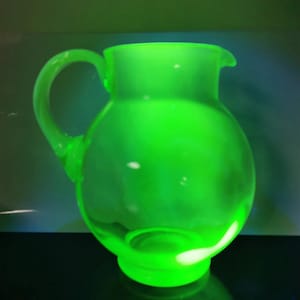 May include: A glowing green glass pitcher with a rounded body, a curved handle, and a pouring spout. The pitcher is made of a translucent material, and the light gives it a bright, vibrant appearance. The pitcher is a decorative item.