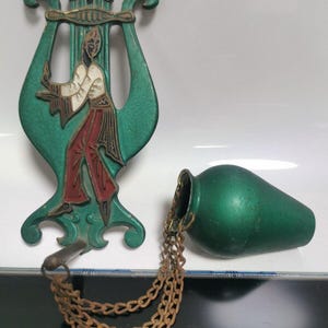 Puede incluir: Un colgante de pared de metal verde en forma de lira con una figura que la toca. Un pequeño jarrón de metal verde cuelga de la lira con una cadena.