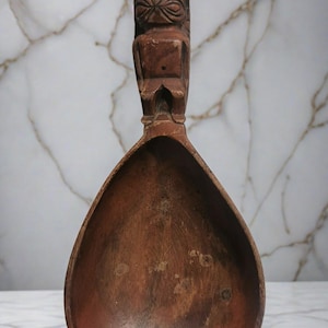 May include: A wooden spoon with a carved handle featuring a Tiki-style figure. The spoon has a large, teardrop-shaped bowl and is made of dark brown wood. The handle has detailed carvings of a face and body.