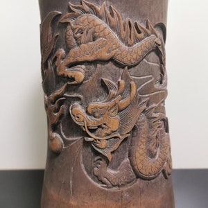 Puede incluir: Un jarrón cilíndrico de madera marrón oscuro con intrincados tallados de dos dragones. Los dragones están tallados en un estilo detallado y realista, con escamas, garras y bigotes.