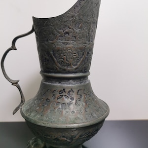 Puede incluir: Una jarra antigua de metal verde con intrincados diseños florales y tres patas de animal. La jarra tiene una base ancha y redondeada y un pico con un asa curva.