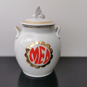 May include: White ceramic honey pot with a gold lid and a bee design. The pot has a red and gold label that says "MED" in a stylized font.