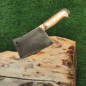 May include: A vintage cleaver with a wooden handle. The blade is made of metal and has a slight curve. The handle is brown and has a light brown colour. The cleaver is sitting on a wooden surface.