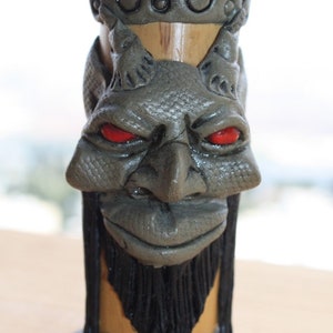 Puede incluir: Una figurita de cerámica gris de una cara con ojos rojos y barba negra. La figurita tiene forma de tapón de botella y tiene una superficie texturizada.