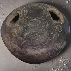 Puede incluir: Un artefacto antiguo redondo, de color gris oscuro, con dos grandes aberturas y diseños geométricos y en espiral grabados. El objeto tiene una apariencia desgastada, lo que sugiere edad e importancia histórica. La superficie muestra signos de desgaste.