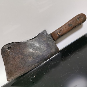 Op de afbeelding: Een vintage hakmes met een houten handvat. Het blad is roestig en heeft een klein gaatje in de buurt van het handvat. Het handvat is bruin en heeft een paar kleine metalen klinknagels.