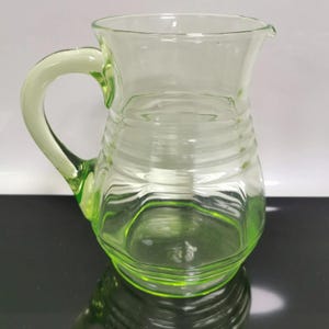May include: A clear, green-tinted glass pitcher with a curved handle and a spout. The pitcher has horizontal ridges around the body. The handle is a lighter shade of green. The pitcher is sitting on a reflective surface.
