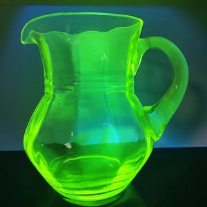 May include: A glowing green glass pitcher with a curved handle and a spout. The pitcher has a rounded body and a scalloped rim. The glass appears to be translucent, with light reflecting off its surface.