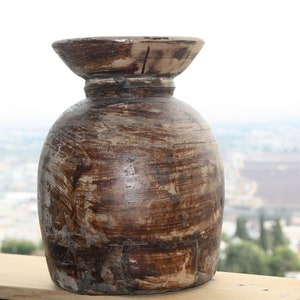 Puede incluir: Un jarrón de madera rústico con un cuerpo ancho y redondeado y una parte superior acampanada. El jarrón está hecho de madera marrón oscuro con vetas visibles y un acabado envejecido. El jarrón se encuentra sobre una superficie de madera.