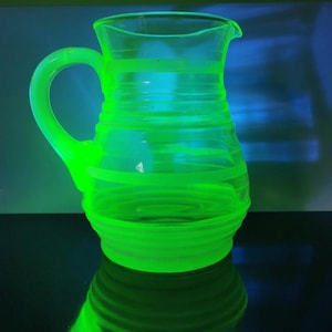 May include: A glowing green glass pitcher with a handle and a spout. The pitcher has horizontal, spiraling lines around its body. The glass is transparent, and the pitcher is reflecting on a dark surface.