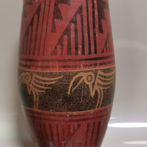 May include: A red clay vase with black geometric designs and bird motifs. The vase is cylindrical in shape and has a smooth, matte finish.