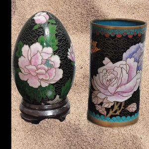 Könnte beinhalten: Zwei Cloisonné-Vasen mit Blumendesigns. Die Vase links ist eiförmig und steht auf einem Holzsockel. Die Vase rechts ist zylindrisch und hat ein blaues und weißes Blumendesign. Beide Vasen haben einen schwarzen Hintergrund mit goldenen Akzenten.