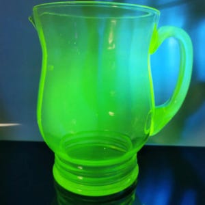 May include: A vibrant, neon green glass pitcher with a curved handle and a spout. The pitcher has a rounded body and a base with concentric rings. The glass appears to be transparent, allowing light to pass through.