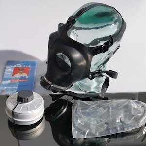 Pode incluir: Uma máscara de gás preta com uma viseira de plástico transparente e um filtro de prata. A máscara está em um frasco de vidro e tem uma correia preta. O filtro está em uma superfície preta. A máscara faz parte de um kit de máscara de gás.