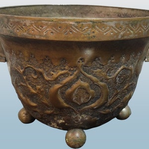 May include: An antique bronze planter with intricate floral designs and three rounded feet. The planter has two handles on the sides.