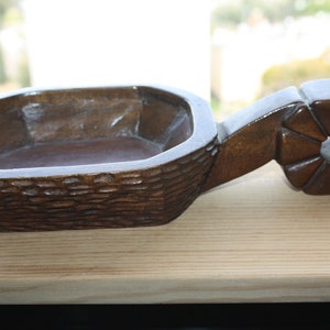 Op de afbeelding: Een donkerbruine houten kom met een gegraveerd handvat. Het handvat is gevormd als een bloem met een klein gaatje in het midden. De kom is gemaakt van ruw, getextureerd hout.