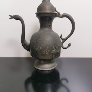 Puede incluir: Una tetera antigua de color plateado con un diseño ornamentado y un pico largo y curvo. La tetera tiene una tapa con un pomo y una asa.