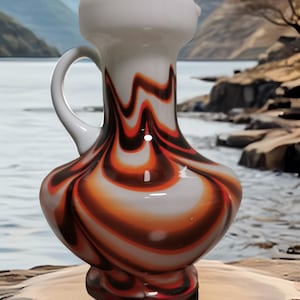 Op de afbeelding: Een decoratieve glazen kan met een witte handgreep en tuit. De body heeft een wervelend patroon van oranje, bruin en wit. De kan heeft een bolvormige basis en een cilindrische hals. De achtergrond toont een meer en bergen.