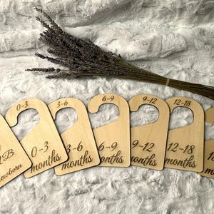 May include: Set of six wooden hangers with size labels for newborn to 18-24 months. The hangers are natural wood with black lettering.