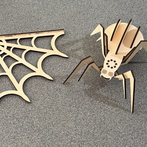 May include: A wooden spider and a spider web corner piece. The spider has a round body with legs and a detailed face. The web is a corner piece with a classic web design. Both are made of light-colored wood.