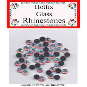May include: A pile of iridescent, round, glass rhinestones with black adhesive backs. The rhinestones are scattered on a white background. The text "#YG-101AB CRYSTAL AB(IRIDESCENT)" is printed below the rhinestones.