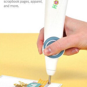 May include: A white Hotee rhinestone setter tool with a blue circular grip section. The tool features a metal tip applying heat-transfer rhinestones to a paper card. Text on the tool reads Hotee. Text on the image reads: Use the rhinestone setter to add individual heat-transfer rhinestones to cards, scrapbook pages, apparel, and more.