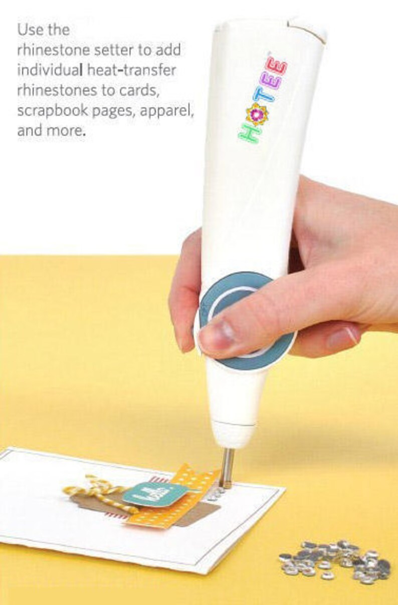 May include: A white Hotee rhinestone setter tool with a blue circular grip section. The tool features a metal tip applying heat-transfer rhinestones to a paper card. Text on the tool reads Hotee. Text on the image reads: Use the rhinestone setter to add individual heat-transfer rhinestones to cards, scrapbook pages, apparel, and more.
