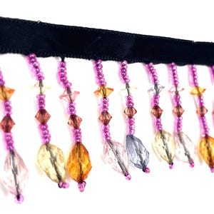 May include: A black ribbon trim featuring hanging beaded fringe. Each strand consists of small pink seed beads interspersed with larger, faceted crystal-like beads in shades of amber, yellow, clear, and light blue, creating a decorative border.